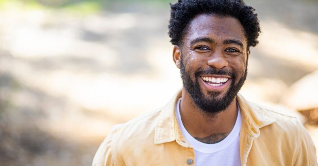 Man with a beard wearing a yellow button up shirt smiling while out in nature