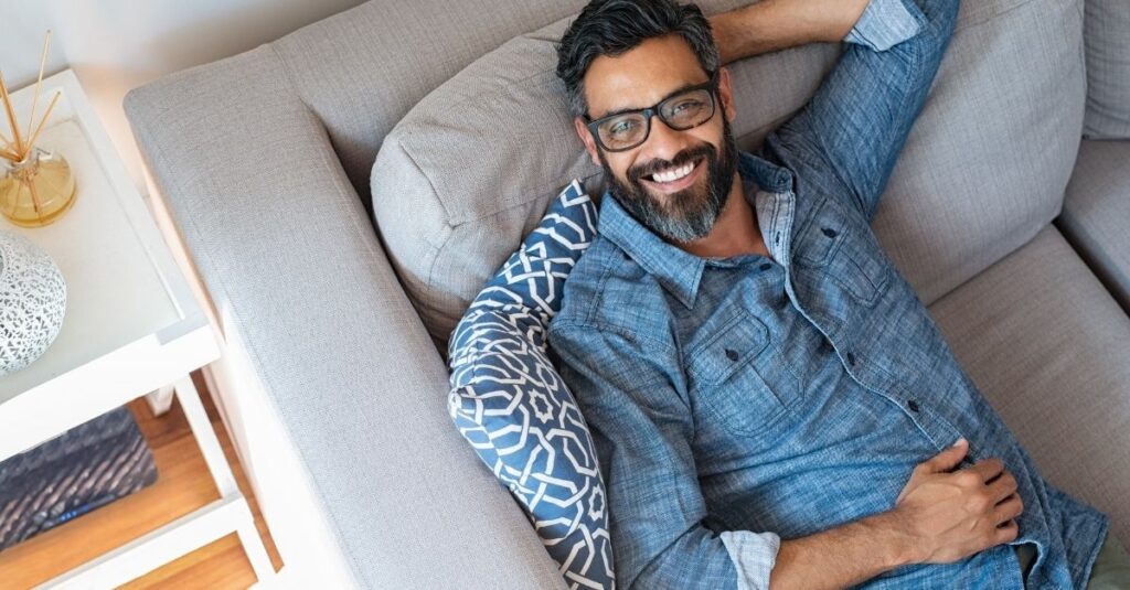 Man relaxing on couch wearing glasses in a blue button up shirt smiling.