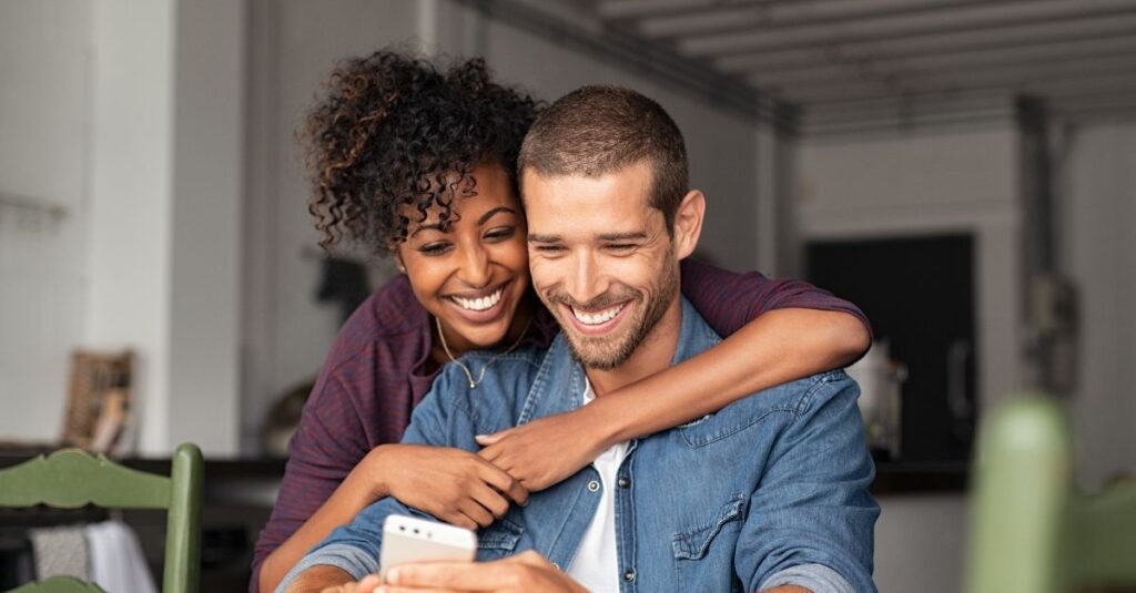A happy couple laugh together while watching a video on their cell phone.