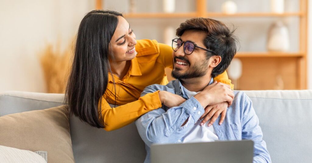 Smiling couple sitting together on a couch, sharing a warm embrace and looking into each other’s eyes, symbolizing emotional connection and renewed intimacy in a long-term relationship.