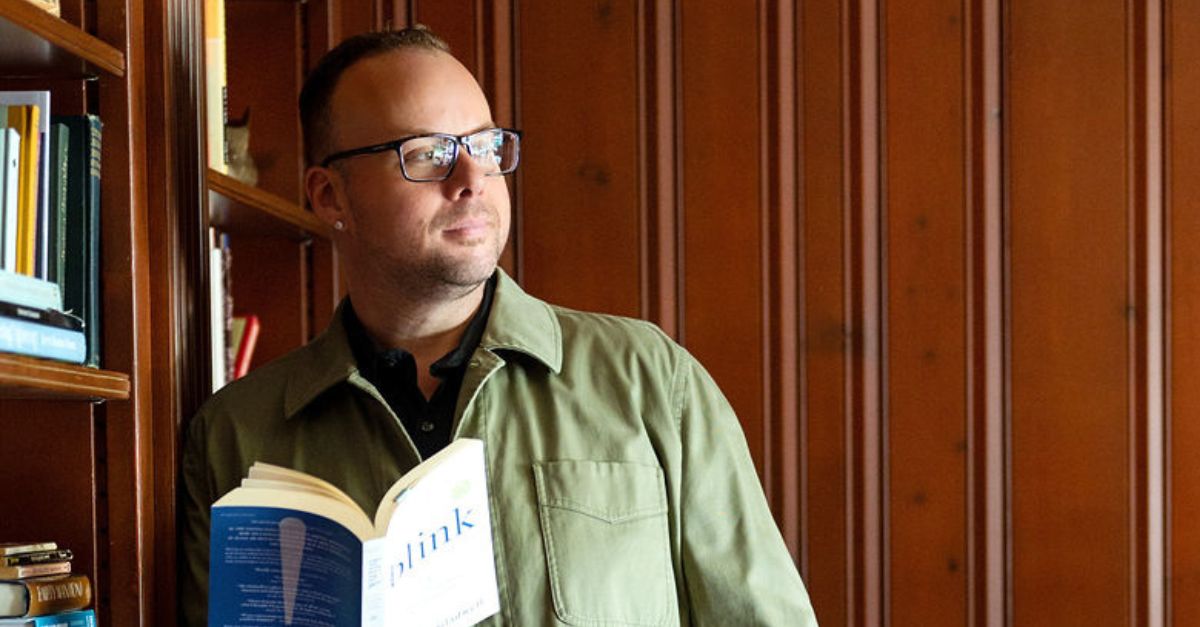 Dr. Mike leans up against a bookshelf while holding a book and looking off into the distance.