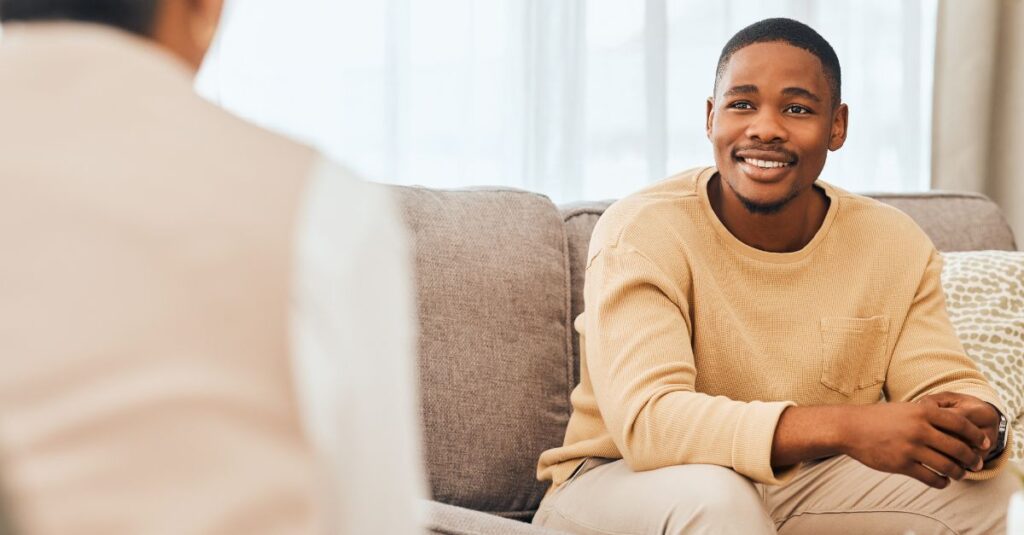 Smiling young man sitting on a sofa, looking relaxed and confident during a therapy session, symbolizing positive outcomes from sex therapy.