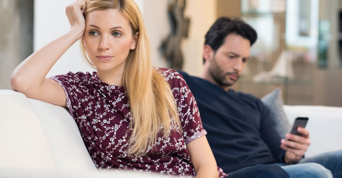 Woman looking upset on the couch while her partner focuses on his phone, symbolizing digital disconnection in relationships.