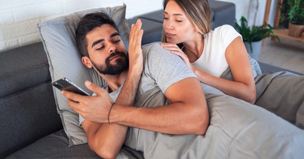 Man in bed focused on his phone while dismissing his partner’s attempt for closeness, representing digital distraction in intimacy.