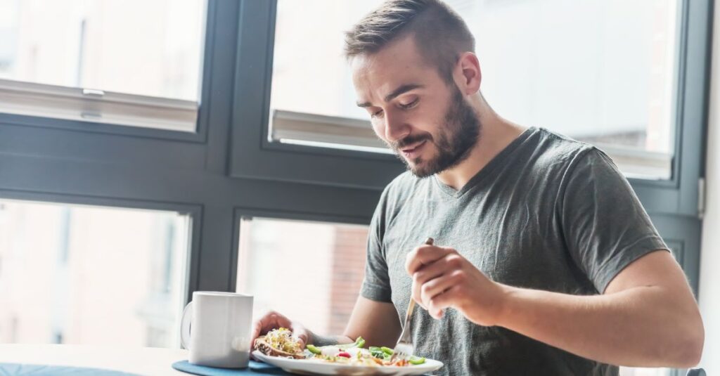 Man sitting at a table by a window, eating a healthy meal with vegetables, representing lifestyle choices that support male fertility and overall wellness.