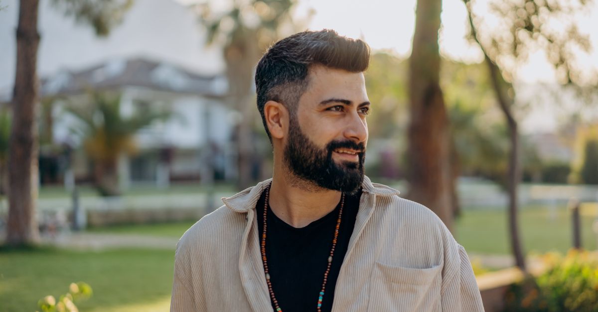 Confident man with a beard standing outdoors, looking to the side with a thoughtful expression.