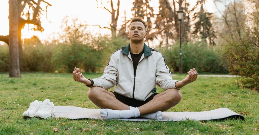 Young man meditating cross-legged on a yoga mat in the park, eyes closed and hands resting on knees in a relaxed pose.