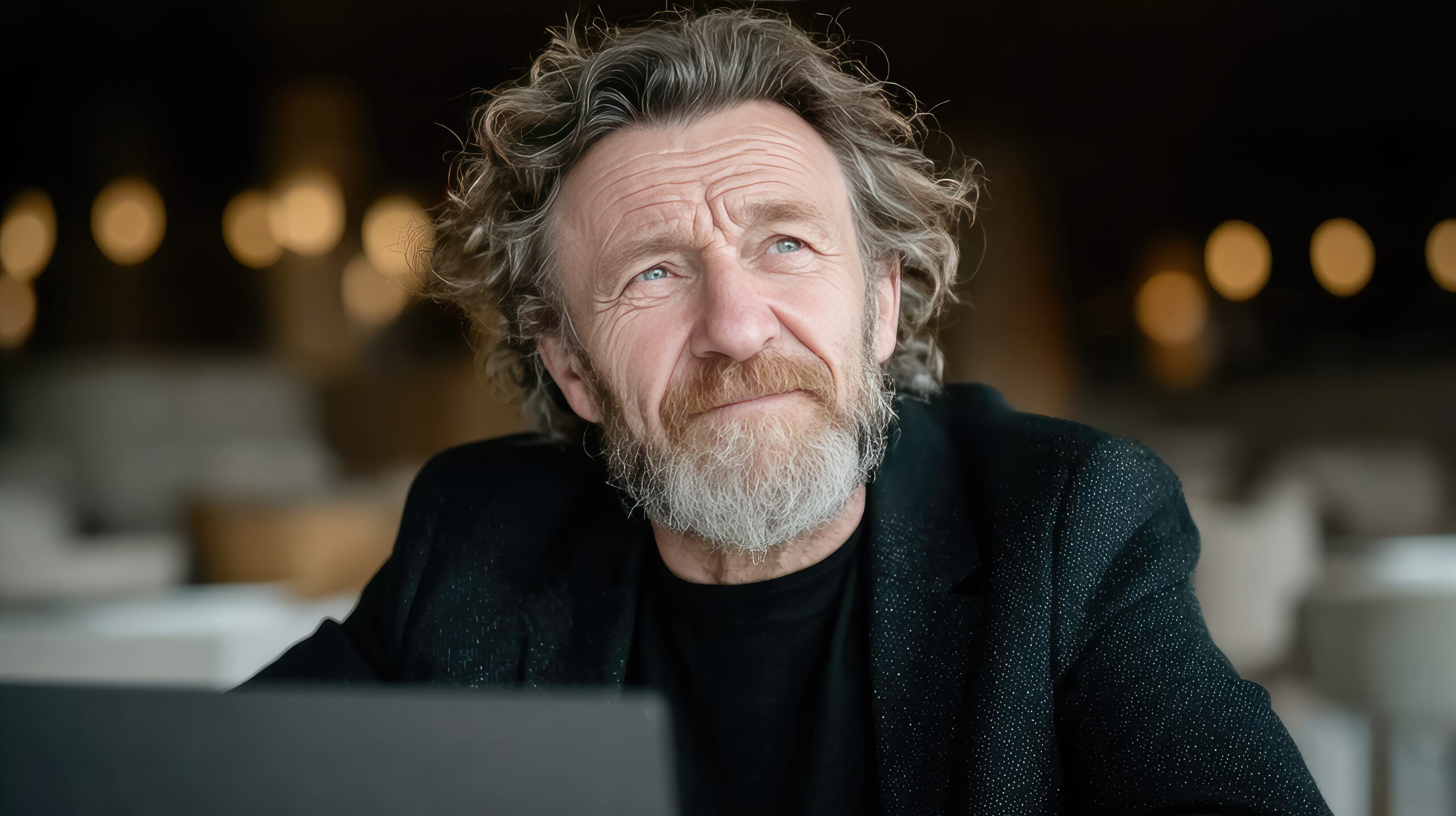 Man with contemplative expression sits at a coffee shop with his laptop open.