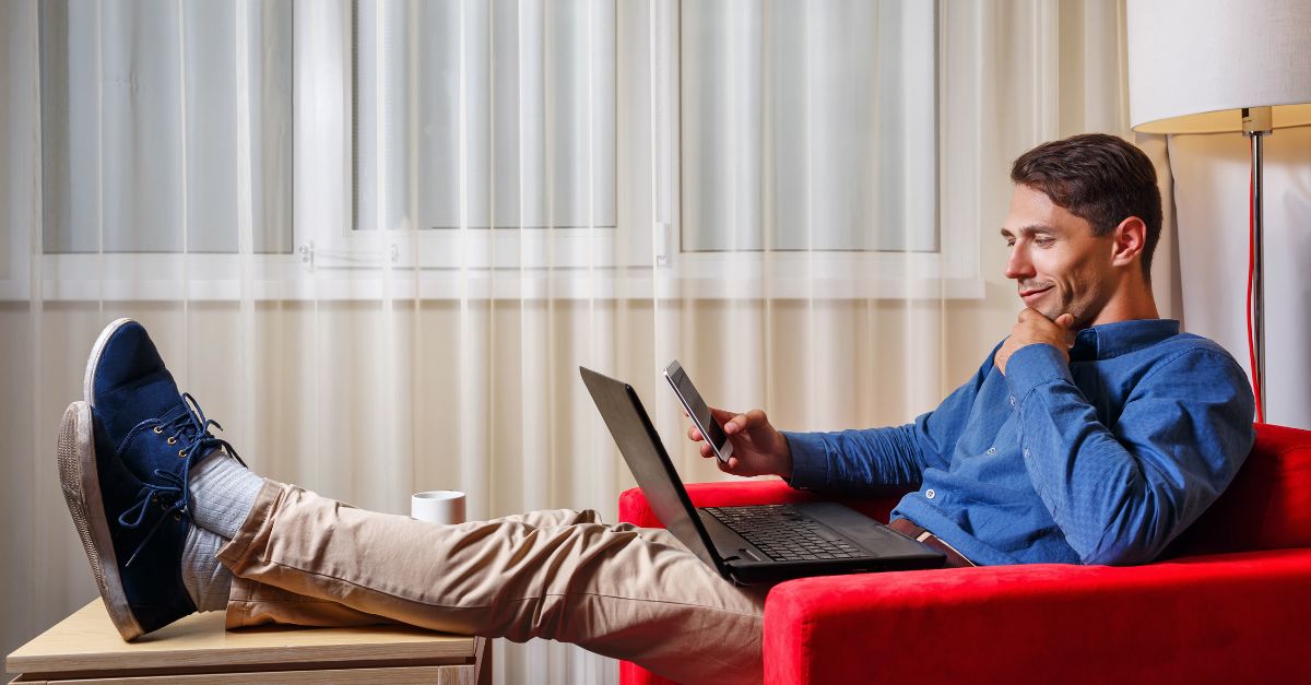 Man relaxing with his feet propped up, a laptop in his lap, and his cell phone in his hand.