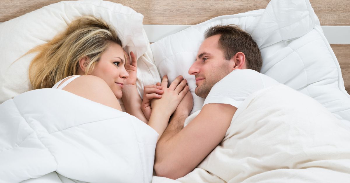 Couple laying in bed holding hands, looking in each other's eyes.