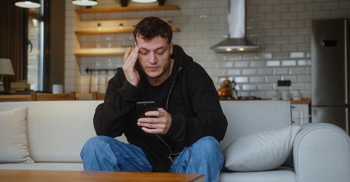 Exhausted man sits on a couch mindlessly scrolling on his phone